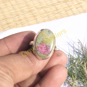 Könnte beinhalten: Ein silberner Ring mit einem ovalen, grün-rosa Edelstein. Der Edelstein hat eine marmorierte Optik mit rosa und grünen Wirbeln.