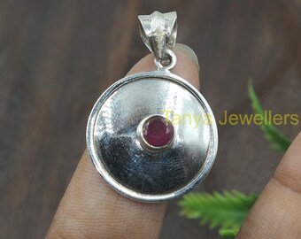 May include: A silver pendant with a round, slightly domed design. The pendant features a small, round, red gemstone in the centre. The pendant is on a finger, and the text "Tanya Jewellers" is visible in the background.