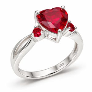 May include: A silver ring featuring a large heart-shaped red gemstone in the center, flanked by two smaller round red gemstones on each side. The band is polished silver and includes a clear 925 hallmark stamp on the inner surface of the ring.