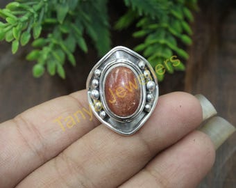 May include: A silver ring with a large, oval, reddish-brown gemstone. The ring has a decorative border with small silver and gold accents. The ring is set on a hand, with green foliage in the background.