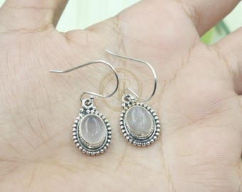 May include: A pair of silver drop earrings featuring oval, pale pink gemstones. Each earring has a beaded border and a simple hook closure. The earrings are displayed on a light-colored surface.