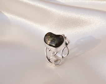 May include: A silver ring with a heart-shaped, dark, iridescent stone. The ring has an open, artistic design with swirling metalwork around the band. The stone has a subtle, reflective surface.