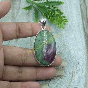 Puede incluir: Un colgante de piedra ovalada verde y rosa con un engaste de plata. El colgante está en una mano.