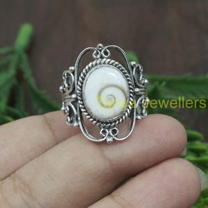 May include: A silver ring with an oval-shaped white shell inlay. The ring has a delicate, ornate design with a rope-like border around the shell.
