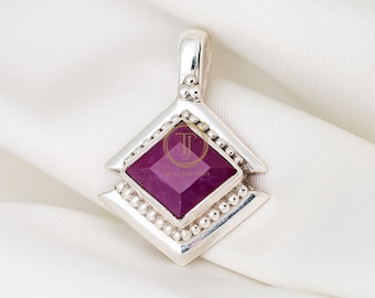 May include: A silver pendant featuring a square, deep magenta gemstone set in a diamond-shaped frame. The frame is decorated with small, round beads and a loop for a chain. The pendant is shown on a white, textured background.