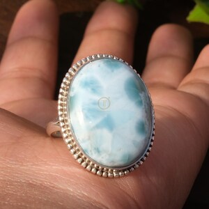 May include: Oval-shaped ring featuring a large, light blue gemstone with a unique pattern. The gemstone is set in a silver band with a beaded border, creating a vintage aesthetic. The ring is displayed on a hand.