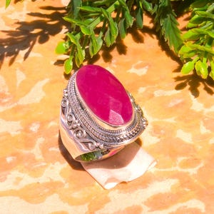 Puede incluir: Un anillo de plata con una piedra preciosa rosa facetada de forma ovalada engastada en una banda de plata decorativa. La banda tiene un diseño de cuerda trenzada y detalles florales intrincados. El anillo está marcado con "925" en el interior.