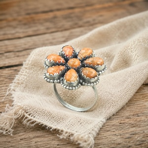 May include: A silver ring featuring a cluster of orange-toned stones arranged in a floral pattern. The stones are set in a scalloped bezel setting. The ring is displayed on a beige fabric, set against a wooden background.