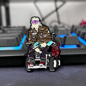May include: An enamel pin showing a person in a wheelchair, wearing a brown jacket and sunglasses. The pin has a red backpack and black wheels. The person has white hair and is seated, holding a blue object.