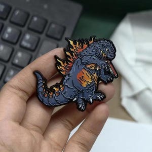 Puede incluir: Un pin de esmalte metálico que muestra un monstruo azul oscuro estilizado con aletas dorsales y marcas de color naranja y amarillo en forma de llama, representado en una pose dinámica con garras y dientes afilados, con un borde de metal negro.