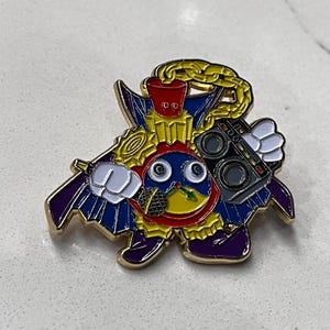 May include: A gold-coloured enamel pin featuring a cartoon character with a yellow face, red hat, and blue cape. The character is holding a microphone and a boombox. The pin has a gold chain around its neck.