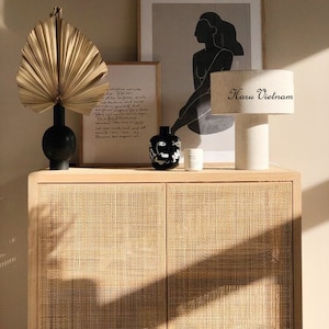 May include: A light wood cabinet with two doors and a woven rattan front. The cabinet is decorated with a dried palm leaf, a black vase, a black and white ceramic vase, a white candle, and a white lamp with a black and white text label that reads "Haru Vietnam".