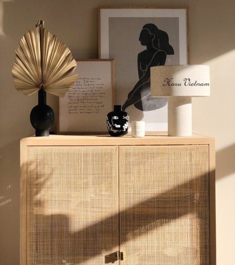 May include: A light wood cabinet with two doors and a woven rattan front. The cabinet is decorated with a dried palm leaf, a black vase, a black and white ceramic vase, a white candle, and a white lamp with a black and white text label that reads "Haru Vietnam".