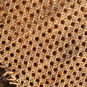 May include: Close-up of a woven rattan material with a repeating pattern of diamond-shaped openings. The natural-colored rattan is tightly woven, creating a textured surface. The image is lit with warm light, highlighting the intricate details of the weave.