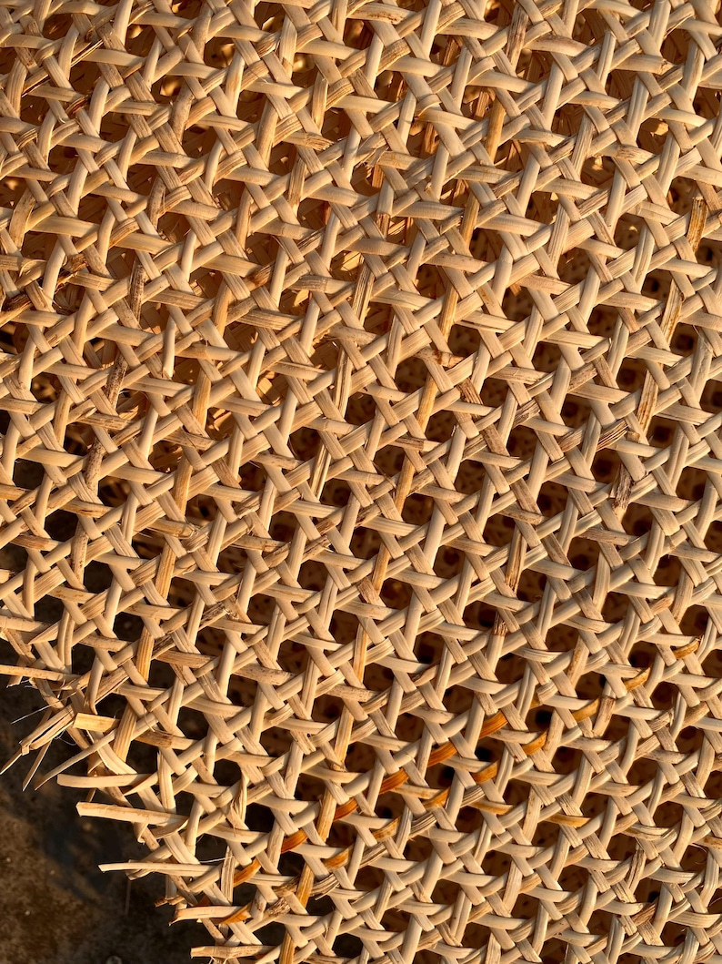 May include: Close-up of a woven rattan material with a repeating pattern of diamond-shaped openings. The natural-colored rattan is tightly woven, creating a textured surface. The image is lit with warm light, highlighting the intricate details of the weave.