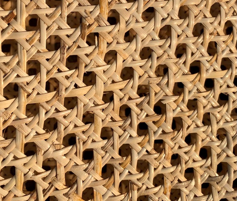 May include: Close-up of a woven rattan material. The natural-toned material is intricately woven in a repeating pattern, creating a textured surface. The weave allows light and shadow to play across the surface, highlighting the detailed craftsmanship.
