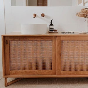 May include: A wooden bathroom vanity with two sliding doors. The doors are made of woven rattan and have a natural wood finish. The vanity has a white porcelain sink and a copper faucet.
