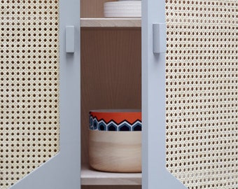 May include: A light blue cabinet with two doors. The doors have a woven cane pattern and are open to reveal shelves. The shelves are empty except for a wooden bowl with a colorful design.