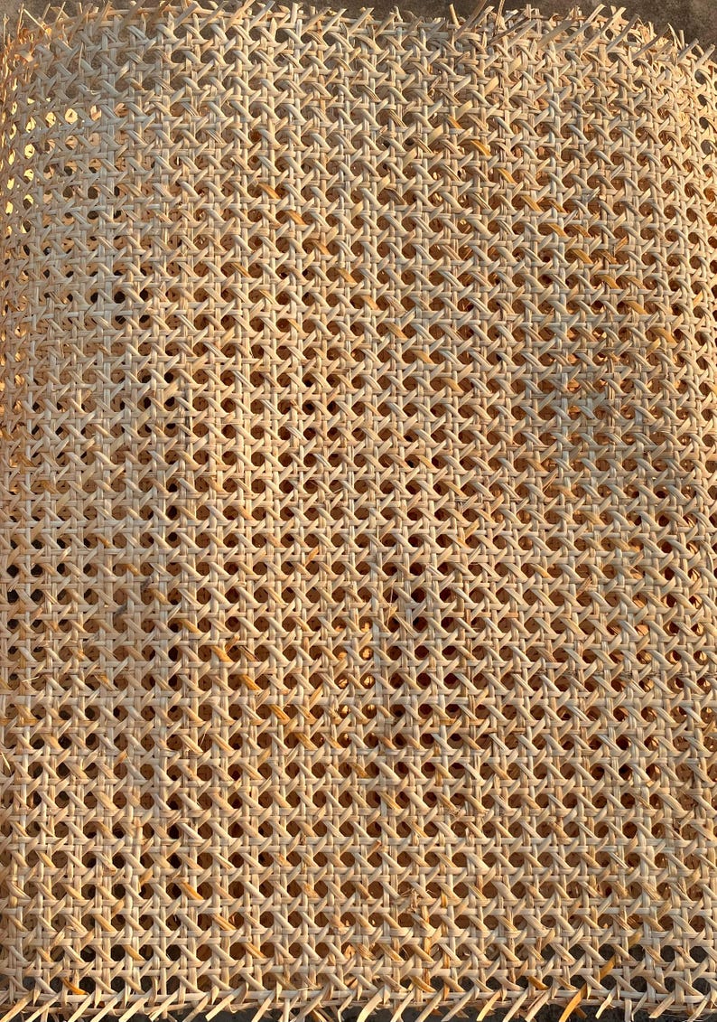 May include: Close-up of a woven cane material with a repeating pattern of small, evenly spaced holes. The natural-colored cane is tightly woven, creating a textured surface. The material is likely used for furniture or decorative purposes.