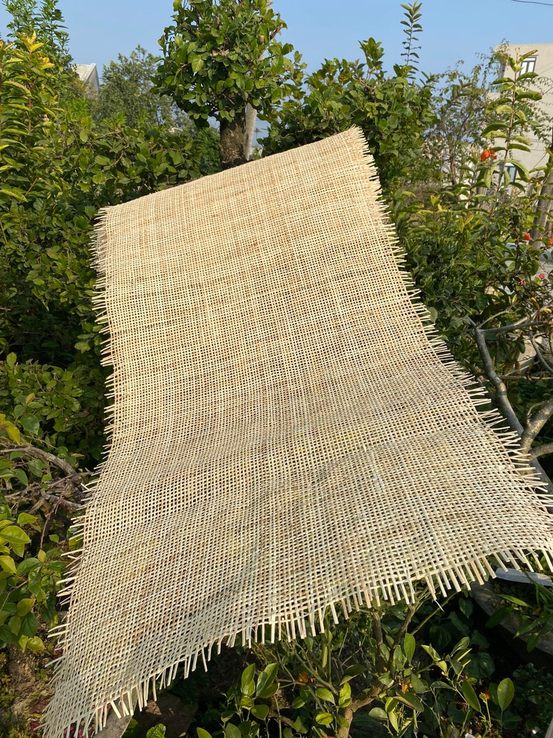 May include: A close-up of a natural woven fabric with a light beige color. The fabric has a grid pattern and is made of thin, woven strands. The fabric is slightly frayed at the edges.