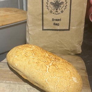 May include: A loaf of sourdough bread on a wooden chopping board. The bread has a golden brown crust and a slightly cracked surface. The bread is next to a brown paper bag with the text "Beeswax Wraps Bread Bag" printed on it.
