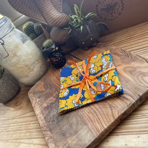 May include: A set of four reusable food wraps featuring a yellow and blue pattern with cartoon characters. The wraps are tied together with an orange ribbon and are sitting on a wooden chopping board.