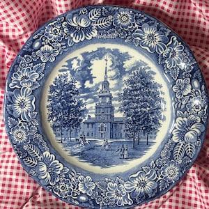 May include: A round ceramic plate featuring a blue and white transferware pattern. The center depicts a historic building with a tall steeple, trees, and figures in a park setting. The rim is decorated with a detailed floral border of blossoms.