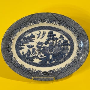 May include: An oval ceramic serving platter featuring a blue and white willow pattern. The central design depicts a landscape with pagodas, trees, a bridge, and two birds in flight. The wide border has a geometric lattice and floral motif.