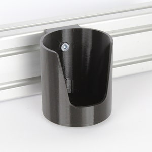 May include: A black plastic cup holder mounted to a silver metal track. The cup holder has a single screw visible on the side.