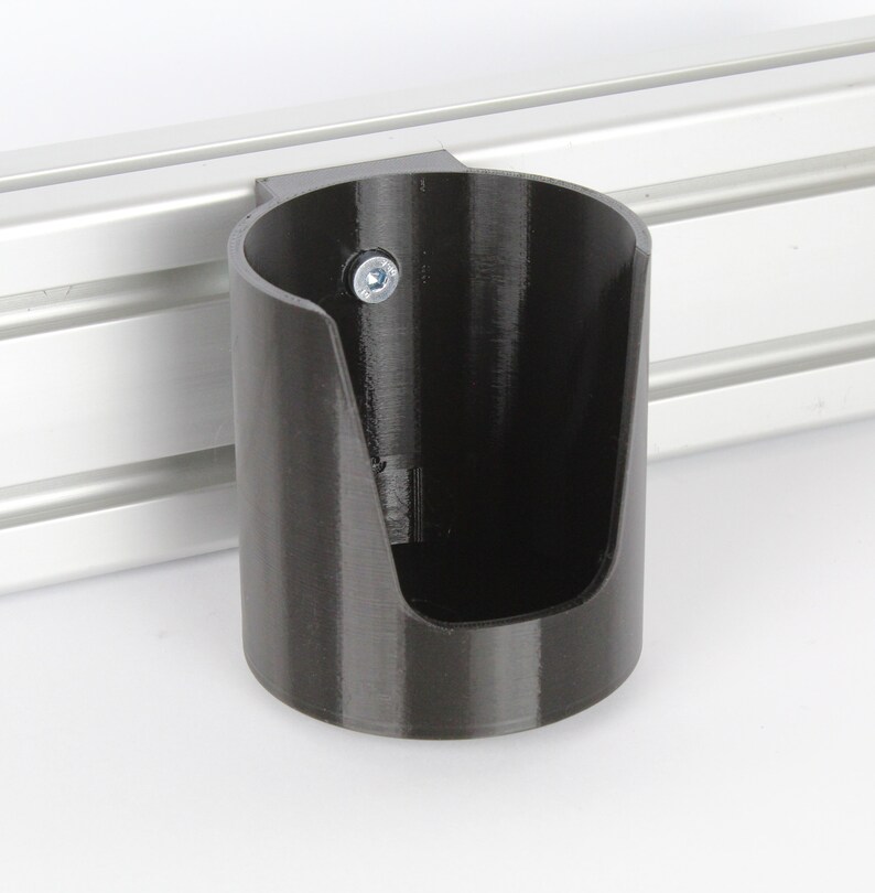 May include: A black plastic cup holder mounted to a silver metal track. The cup holder has a single screw visible on the side.