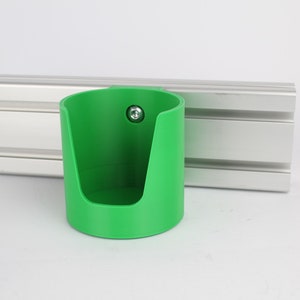 May include: A green plastic cup holder with a screw attached to the side. The holder is mounted on a silver aluminum track.