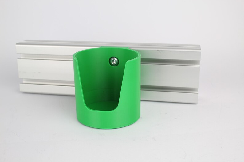 May include: A green plastic cup holder with a screw attached to the side. The holder is mounted on a silver aluminum track.