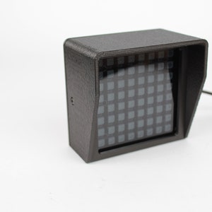 Può includere: Un involucro rettangolare nero stampato in 3D con un piccolo display LED quadrato bianco e nero. L'involucro ha una piccola visiera inclinata nella parte superiore.