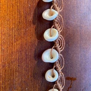 May include: A close-up of a brown macrame cord bracelet with white shell beads. The bracelet is made with a knotting technique and features a series of round, white shell beads.