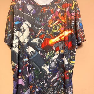 Può includere: Una t-shirt a maniche corte con girocollo caratterizzata da una stampa grafica all-over di vari personaggi robotici in una scena di battaglia. Il design include molteplici figure nei toni del rosso, blu, grigio e giallo, con brillanti esplosioni di energia arancione.