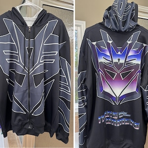 Peut inclure: Sweat-shirt à capuche noir avec fermeture éclair, orné d'un grand logo Transformers blanc et violet sur le dos. Le devant du sweat-shirt présente un logo plus petit, gris et blanc. Le texte "Transformers" est imprimé en blanc sur le dos du sweat-shirt.