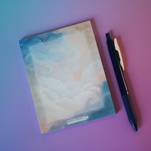 May include: A notepad with a cloud design and the word "MONOTONOUS" in the corner. The notepad has a blue and peach color scheme. A black pen with a white clip rests beside the notepad on a purple surface.
