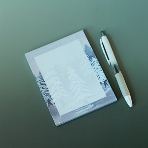 May include: A notepad featuring a winter scene illustration and a white pen with a grey grip. The notepad has a snowy forest design with the text "MONOTONOUS" at the bottom. The pen is placed beside the notepad on a green surface.