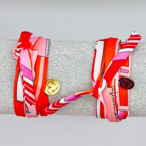 Könnte beinhalten: Zwei bunte Seidenschal-Armbänder. Die Armbänder weisen rote, rosa und orangefarbene Muster mit gold- und silberfarbenen Charms auf. Die Armbänder sind um einen grauen zylindrischen Gegenstand gewickelt.