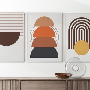 May include: Three framed abstract art prints with geometric shapes in shades of brown, orange, and yellow. The prints are hung on a white wall above a wooden cabinet.