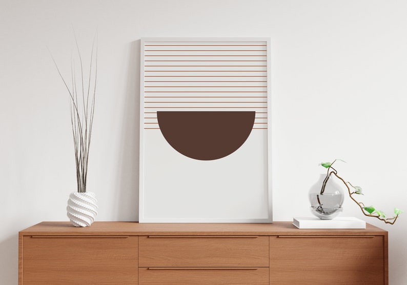May include: A white framed print with a brown half-circle shape and thin horizontal lines in a light brown color. The print is hanging on a white wall above a wooden dresser with two drawers.