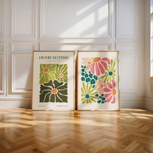 Könnte beinhalten: Zwei gerahmte Kunstdrucke mit Blumenmotiven. Ein Druck zeigt grüne und orangefarbene Blumen mit dem Text "HENRI MATISSE PAPIERS DECOUPES". Der andere Druck hat rosa, orange und blaue Blumen. Beide sind in goldenen Rahmen.