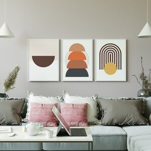 May include: Three abstract geometric wall art prints in a living room setting. The prints feature a brown half-circle, a stack of orange, brown, and gray semi-circles, and a brown arch with a yellow circle.