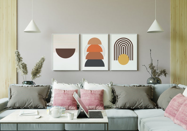 May include: Three abstract geometric wall art prints in a living room setting. The prints feature a brown half-circle, a stack of orange, brown, and gray semi-circles, and a brown arch with a yellow circle.