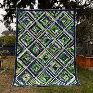 May include: A large, handmade quilt with a geometric diamond pattern. The quilt features a patchwork design with shades of green, blue, white, and gray. The quilt is hanging on a clothesline in a backyard setting.