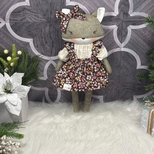 May include: A handmade fox doll with a grey head, white ears, and a floral bow. The doll wears a white blouse and a floral print dress. The doll is standing on a white fluffy surface, with a grey and white patterned background.