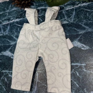 May include: Cream-coloured baby overalls featuring a subtle grey swirl design. The dungarees have frilled straps and a comfortable fit. A small tag is attached. Ideal for a baby shower or a newborn's first outfit.