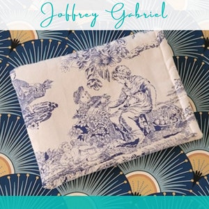 May include: A white fabric pouch with a blue and white toile pattern featuring a pastoral scene with two figures. The pouch has a folded edge and is on a blue and gold patterned background.