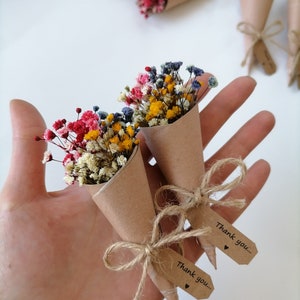 Puede incluir: Pequeños ramos de flores secas de colores en conos de papel marrón, atados con hilo y etiquetas que dicen "Thank you...". Las flores incluyen flores rojas, amarillas, rosas y azules. Varios ramos son visibles.