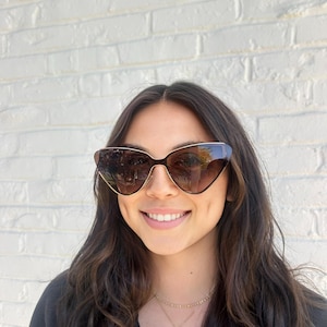 May include: Cat-eye sunglasses with a brown gradient lens and gold-coloured frames. The sunglasses have a sleek, modern design. The image shows a person wearing the sunglasses.